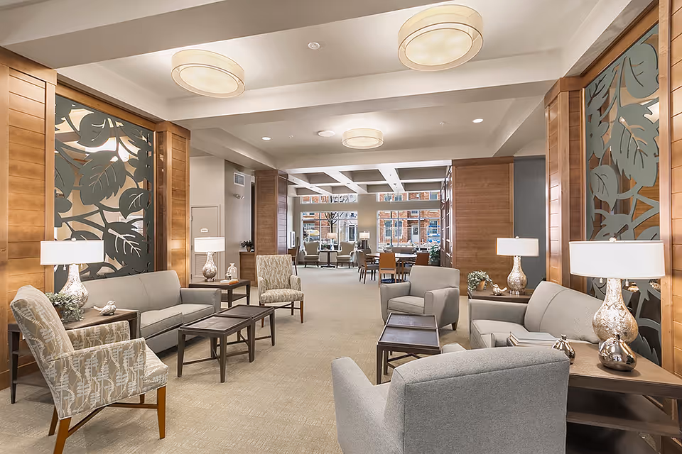Spacious, well-lit senior living lounge with sofas, armchairs, coffee tables, lamps, and decorative wood-paneled partitions.