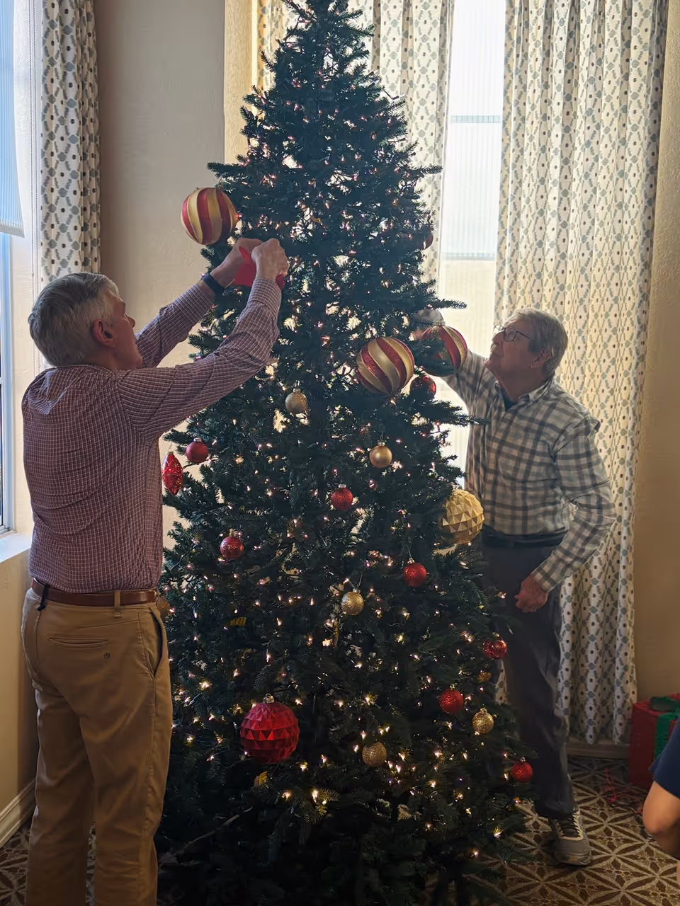 Two people decorating a large Christmas tree in an interior room with patterned curtains and a window.