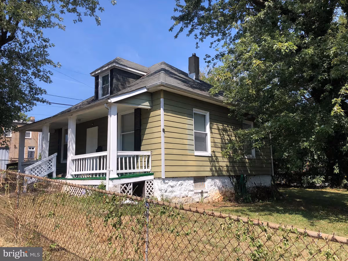 Exterior view of a single-story house with green siding and white trim, featuring a small covered porch with white railings. The house is surrounded by a chain-link fence and trees, under a clear blue sky.