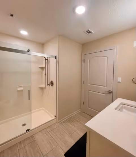 A clean and modern bathroom featuring a glass-enclosed shower with a sliding door, a built-in shelf, and a wall-mounted showerhead. The room has beige walls, a white door with a silver handle, recessed ceiling lights, and a white countertop with an integrated sink. The floor is covered with light-colored tiles, and a black mat is placed near the sink.