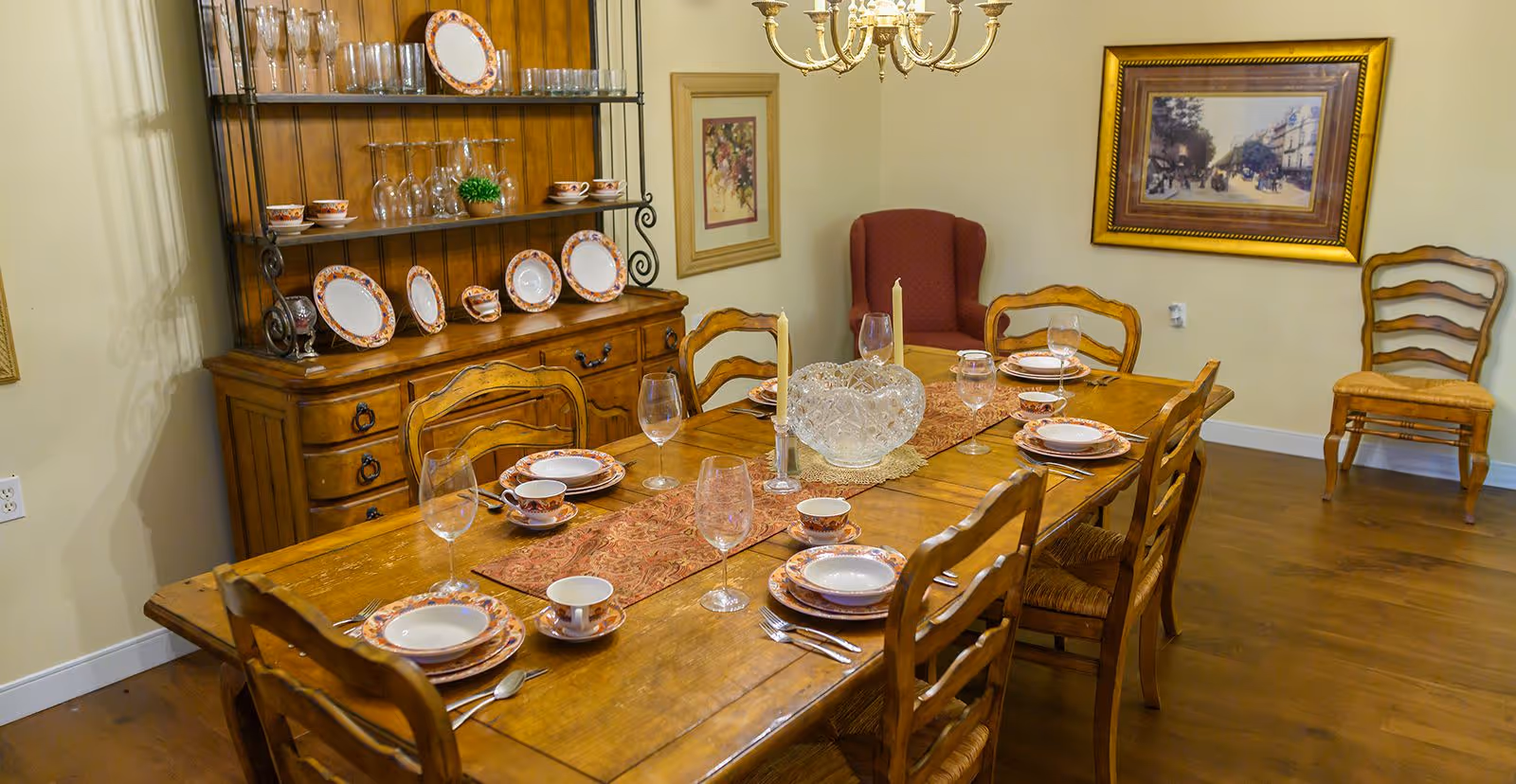 A dining room with a wooden table set for six people with plates, bowls, cups, wine glasses, and silverware. The table has a decorative runner and two tall candles. Behind the table is a wooden hutch displaying matching plates, cups, and glassware. The room has wooden flooring, a chandelier, framed artwork on the walls, and a red upholstered armchair in the corner.