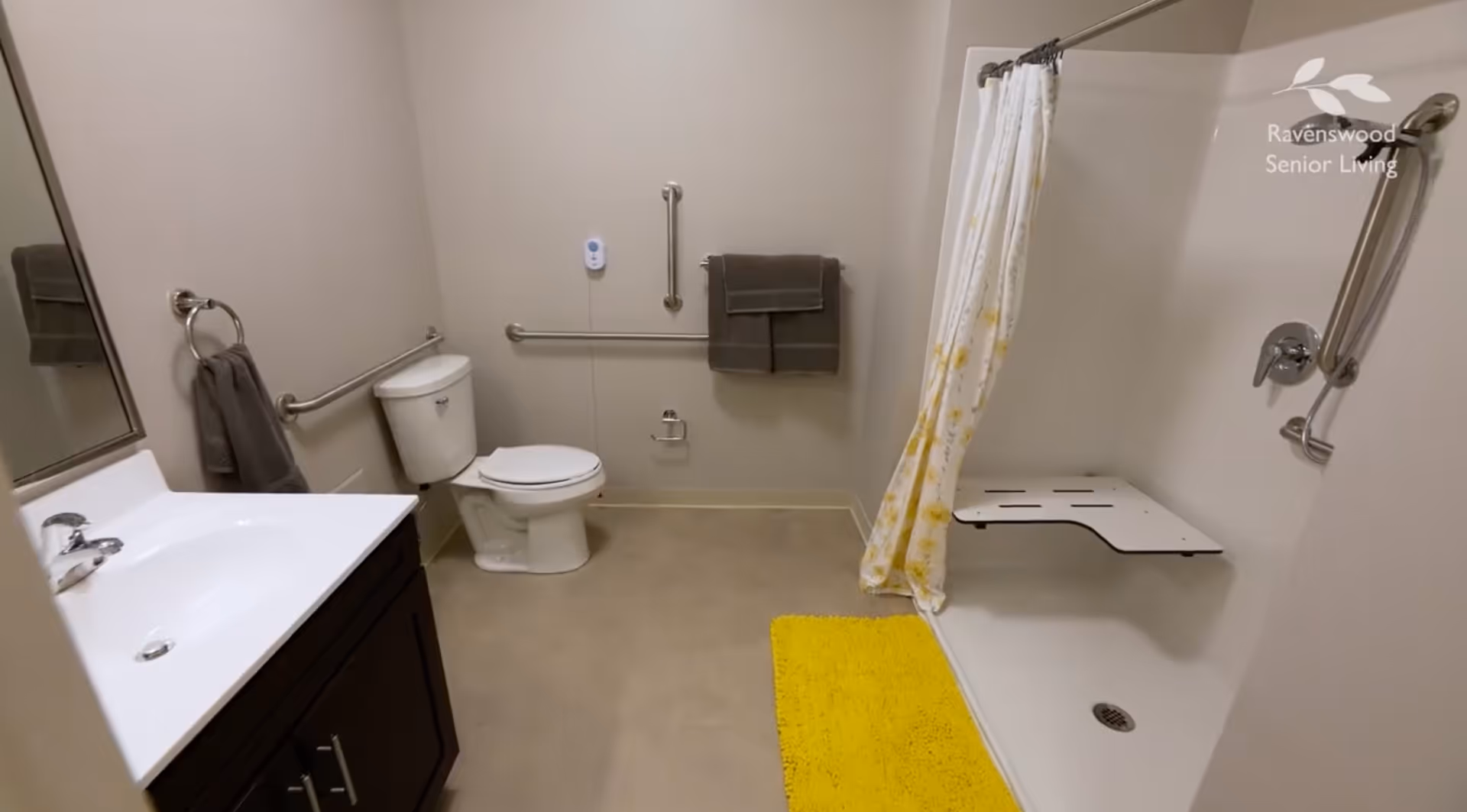 A clean and accessible bathroom with a white toilet, a sink with a dark wooden cabinet, grab bars on the walls, a shower area with a fold-down seat and grab bars, a yellow bath mat, and a shower curtain with a floral pattern. Two gray towels hang on the wall. The wall has a logo that reads Ravenswood Senior Living.