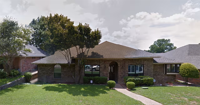 Front exterior view of a single-story brick building with a sloped roof, a central entrance with an arched doorway, several windows, and a well-maintained lawn with trimmed bushes and trees.