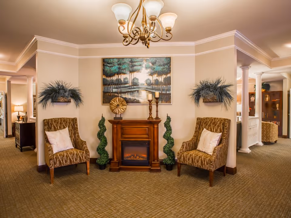 A cozy sitting area in a senior living facility featuring two patterned armchairs with white pillows, a wooden electric fireplace with decorative items on top, two small topiary plants on either side, and a large painting of a forest and river above the fireplace. The space is warmly lit with a chandelier overhead and has beige walls and carpeted floors.