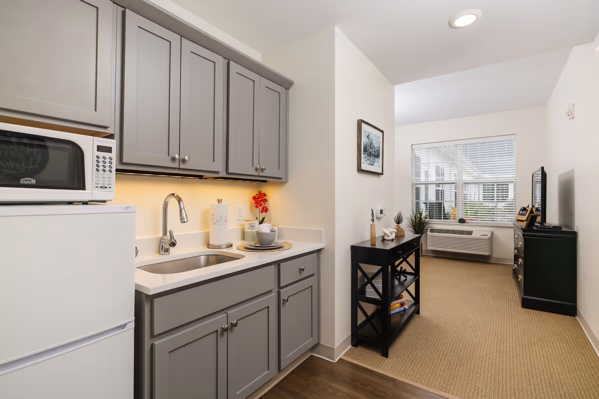 Small kitchenette with gray cabinets, sink, microwave and refrigerator opening into a carpeted living area with a window and air-conditioning unit.