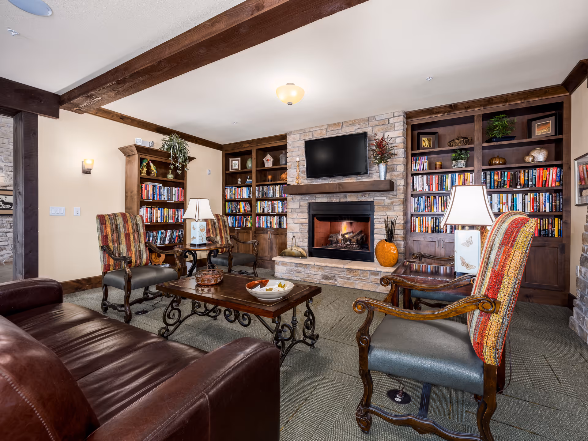 Cozy common room with leather sofa, upholstered chairs, coffee table, built-in bookshelves and a stone fireplace with a mounted TV.