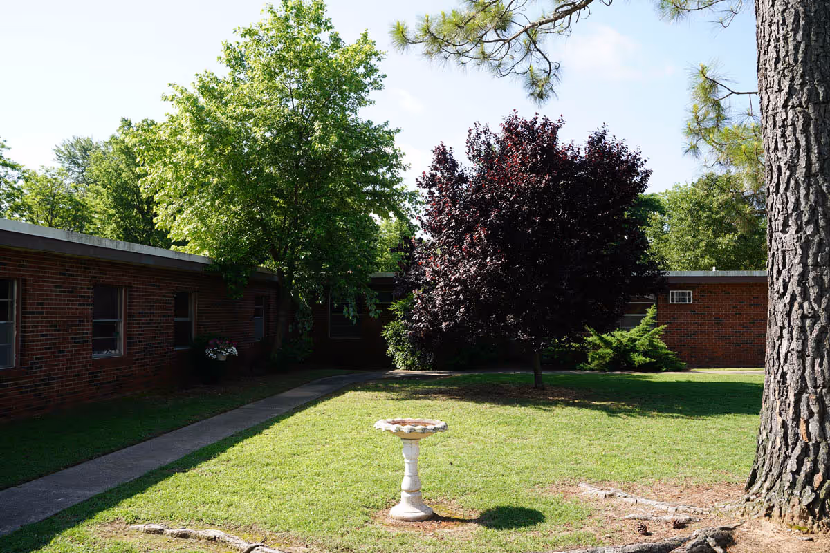 Courtyard of a single-story brick nursing facility with a grassy lawn, trees, a birdbath, and a concrete walkway.