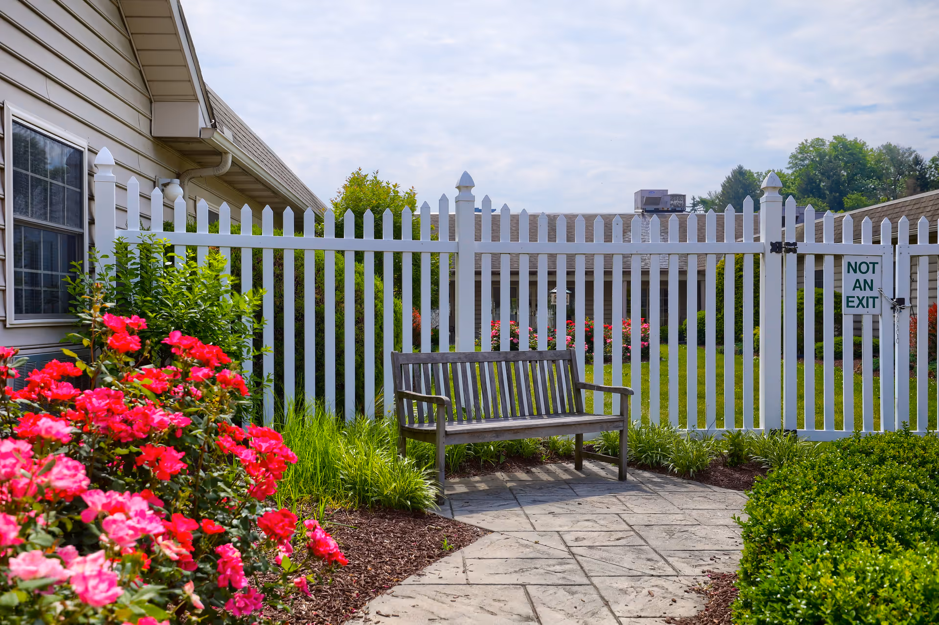 A peaceful outdoor garden area with a wooden bench on a stone pathway, surrounded by vibrant pink flowers, green bushes, and a white picket fence with a sign that reads 'NOT AN EXIT'.
