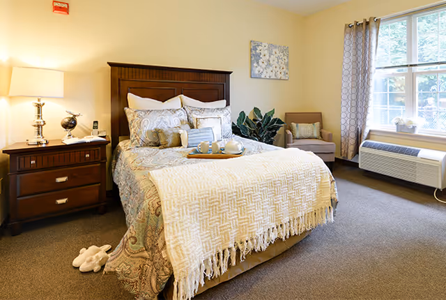Bright, furnished bedroom with a made bed, nightstand, armchair, potted plant and window air conditioner.