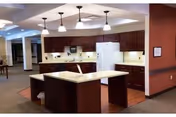 Communal kitchen area with a central island, pendant lights, dark wood cabinets, refrigerator and microwave.