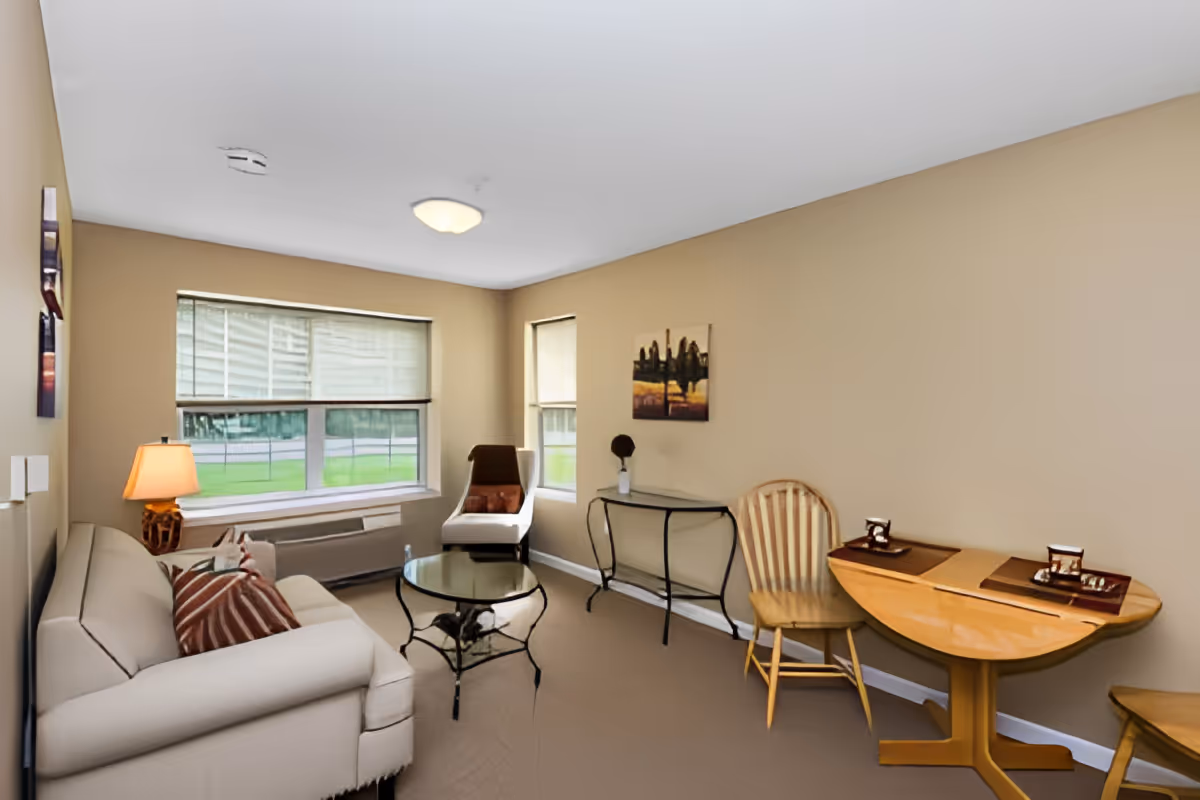 A cozy living room with beige walls and carpet. The room features a white sofa with striped pillows, a glass coffee table with a black metal frame, a wooden chair, and a small wooden drop-leaf table set with placemats and decorative items. There is a window with blinds letting in natural light, a floor lamp with a beige shade, and a small console table with a decorative vase and artwork on the wall.