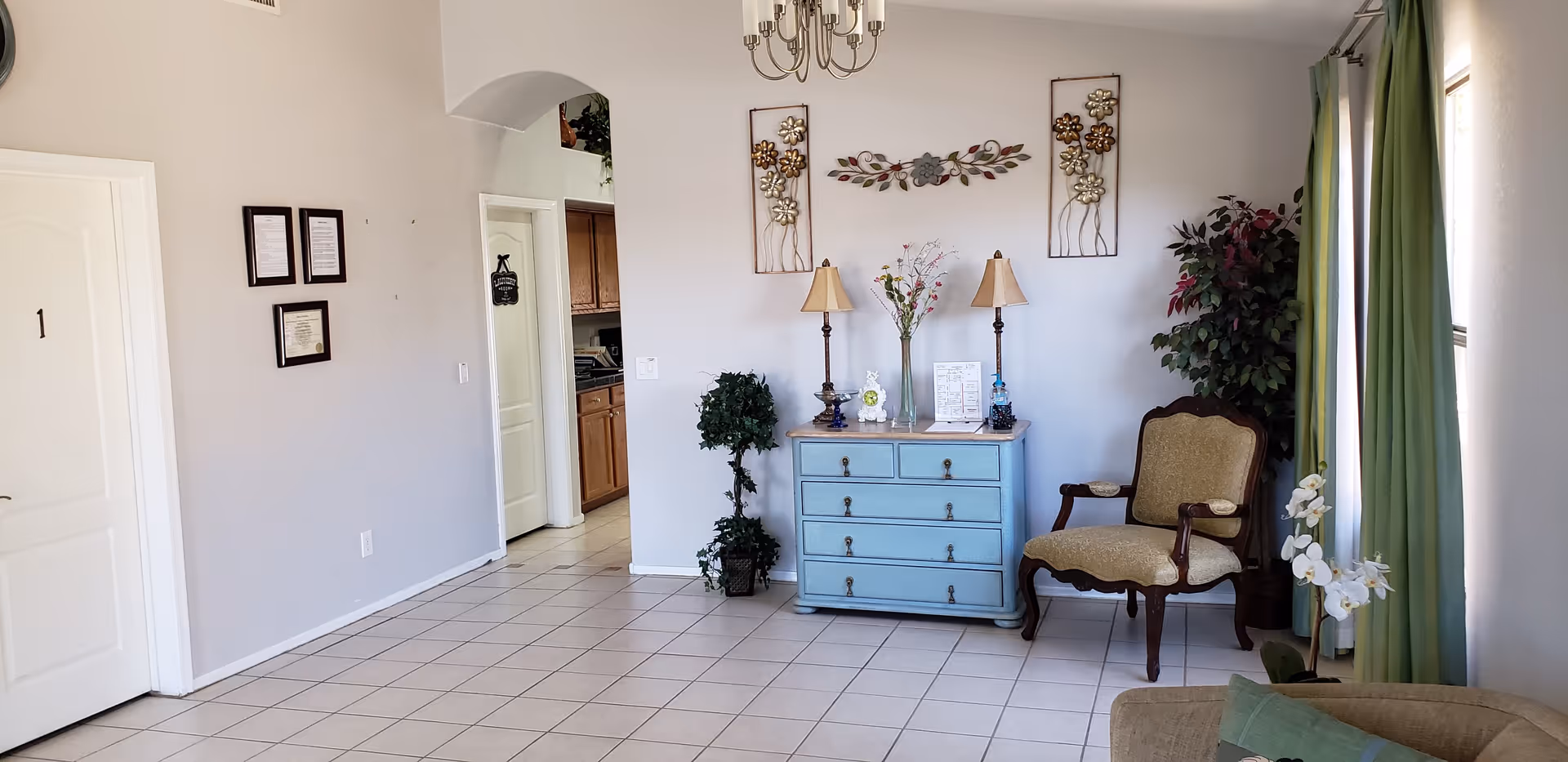 Tile-floored sitting area featuring a blue dresser with lamps, an upholstered armchair, decorative wall art, and a window with green curtains.