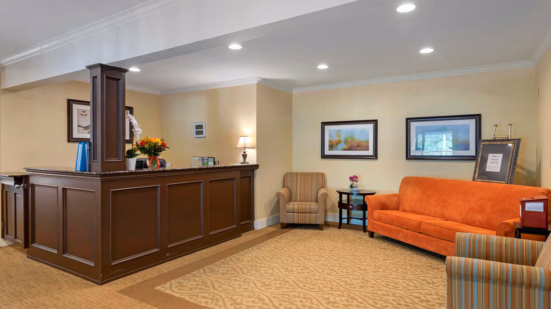 Reception area with a dark wood front desk adorned with a flower vase and office supplies. To the right, there is a seating area with an orange sofa, two striped armchairs, a small round table with a flower vase, and framed artwork on the beige walls. The floor is carpeted with a patterned design, and the ceiling has recessed lighting.