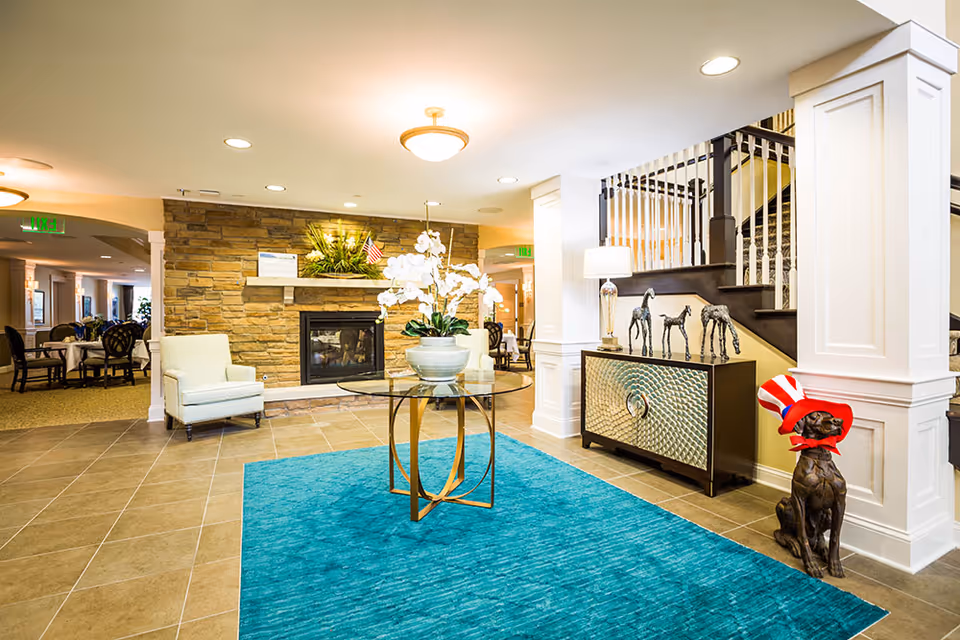 Bright and spacious senior living facility common area with a stone fireplace, a glass table with a white orchid centerpiece on a blue rug, two armchairs, a decorative cabinet with horse sculptures, and a dog statue wearing a red, white, and blue hat near a staircase.