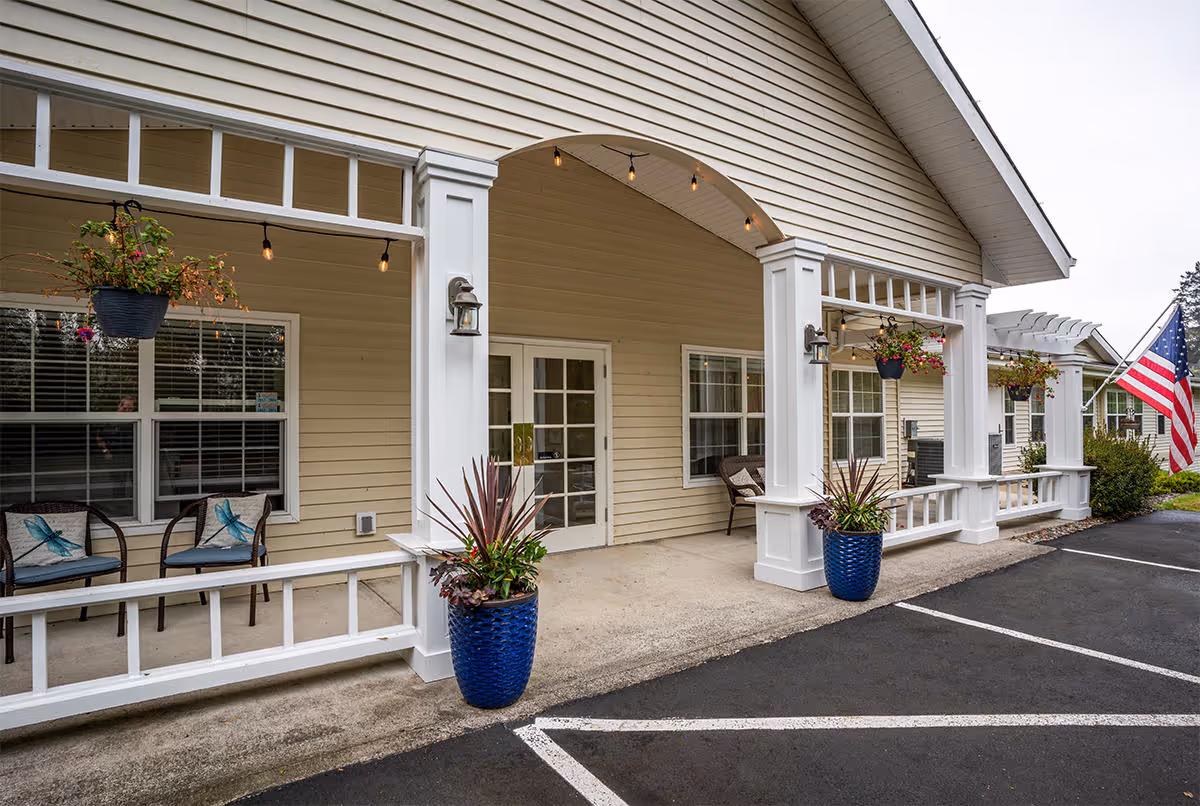 Exterior view of a senior living facility entrance with beige siding, white columns, and a covered porch area. There are hanging flower pots, potted plants in blue containers, chairs with cushions, and an American flag displayed near the parking area.