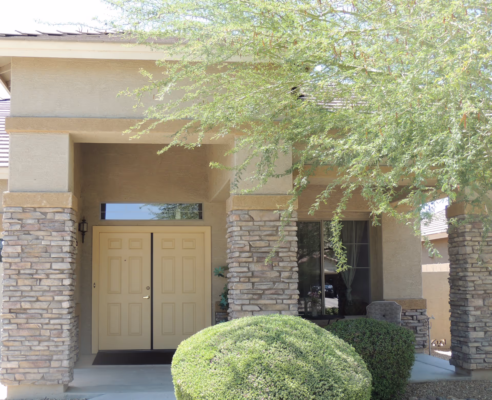 Front entrance of a building with double beige doors, stone pillars, and a small porch area partially shaded by tree branches and surrounded by neatly trimmed bushes.
