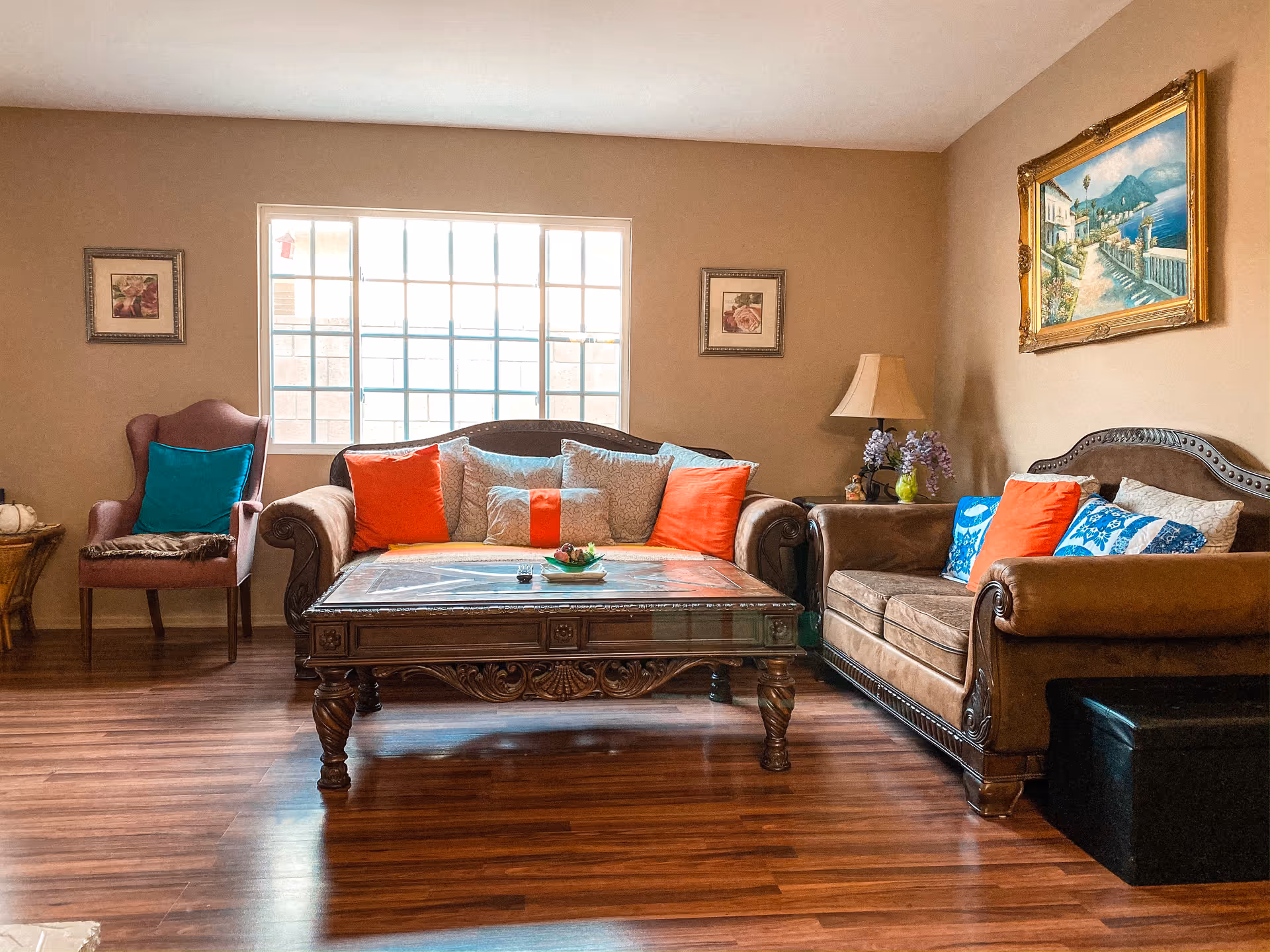 Sunlit living room featuring two ornate brown sofas, a carved coffee table, colorful throw pillows, an armchair, a lamp, and wall art.