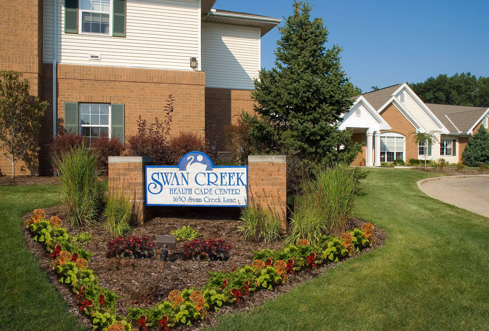 Exterior view of Ohio Living Swan Creek Health Care Center with a landscaped garden featuring green grass, colorful plants, and a sign displaying the facility's name and address in front of a brick and siding building under a clear blue sky.