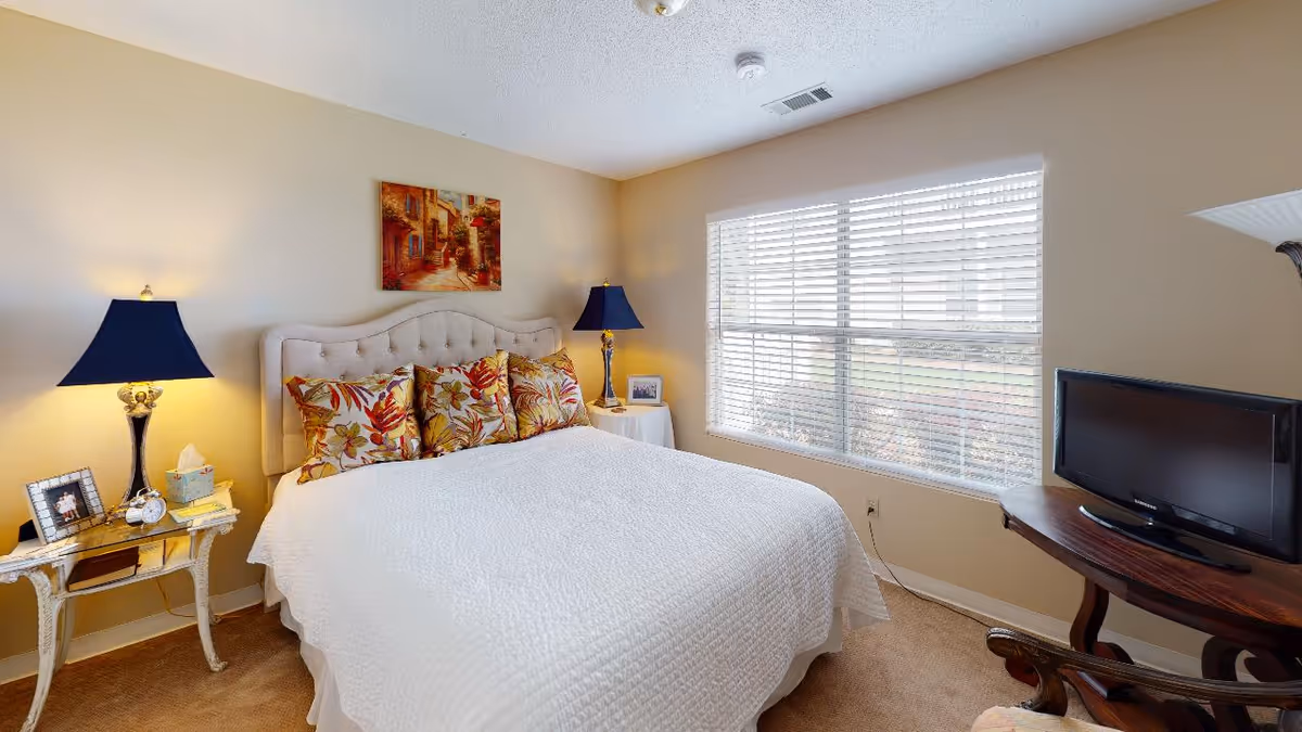 A cozy bedroom with a large bed covered in a white quilt and floral pillows. Two bedside tables each hold a lamp with a dark blue shade, a framed photo, and other small items. A window with white blinds lets in natural light, and a small TV sits on a wooden table near the window. A painting hangs on the beige wall above the bed.
