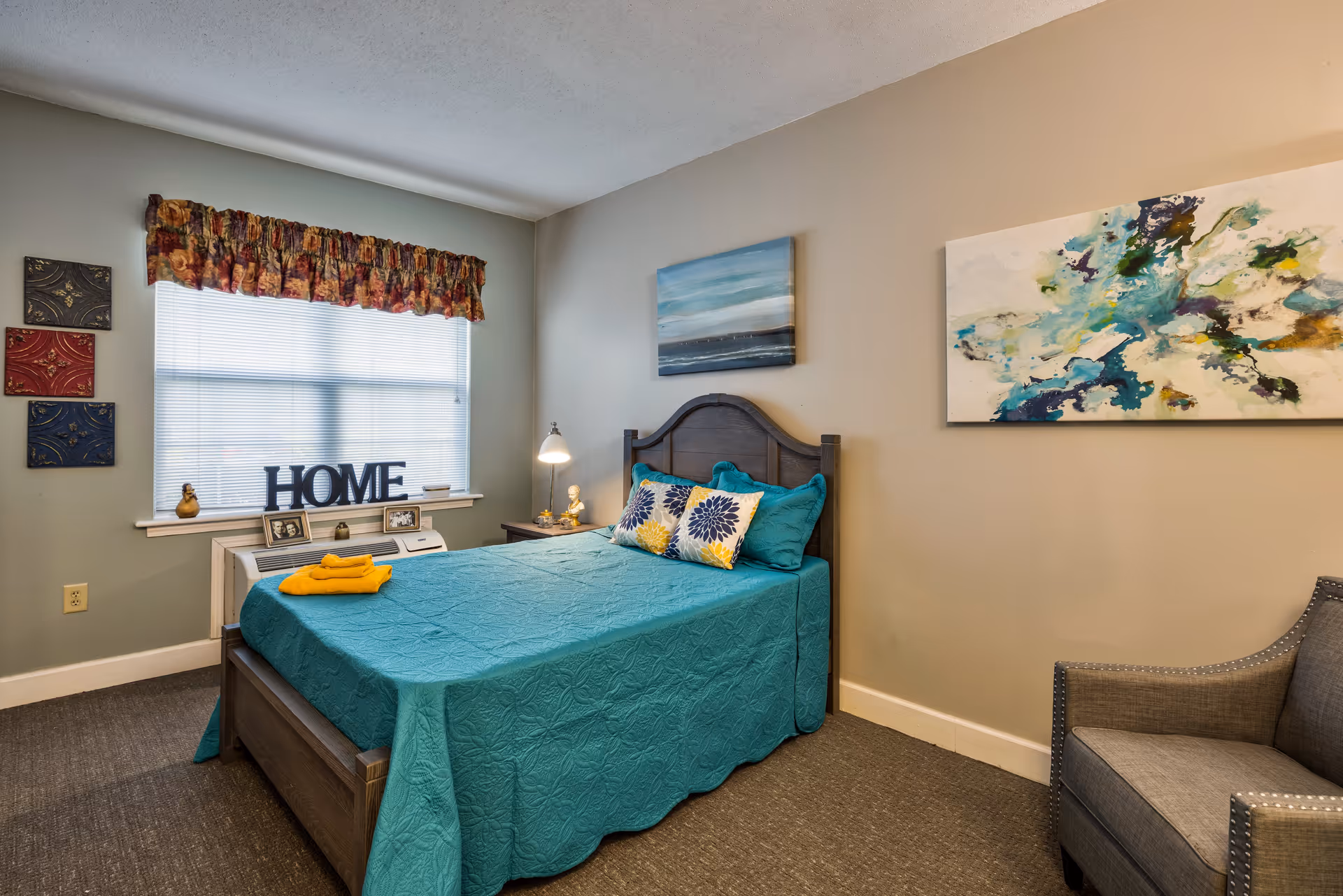 A cozy bedroom with a teal bedspread on a wooden bed frame, two decorative pillows, and a folded yellow towel on the bed. The room has a window with blinds and a floral valance, a small white dresser with framed photos and a decorative HOME sign, a bedside table with a lamp, abstract paintings on the walls, and a gray upholstered armchair.