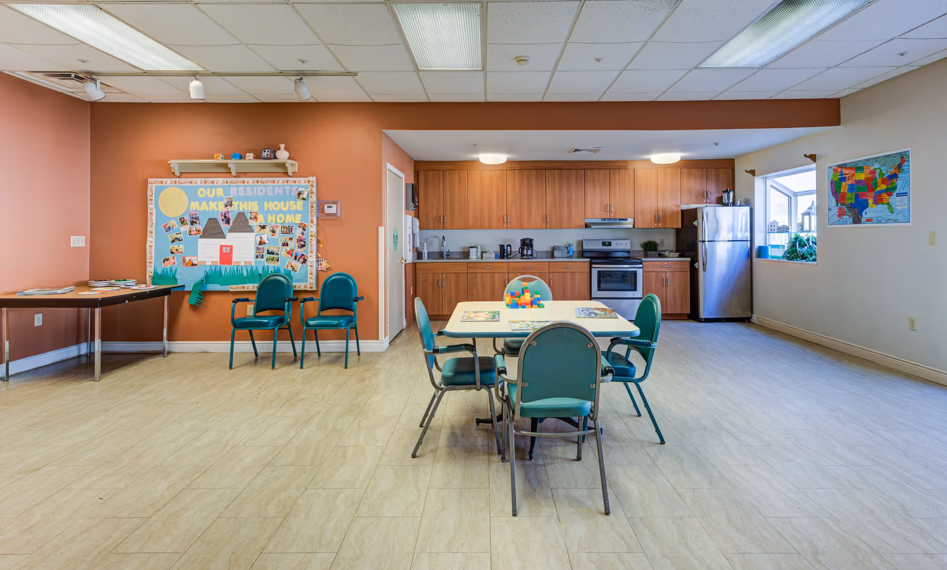 A spacious room with a kitchen area in the background featuring wooden cabinets, a stainless steel refrigerator, stove, and microwave. In the foreground, there is a table with four green chairs around it and some colorful blocks on the table. On the left side, there are two additional green chairs and a bulletin board with the text 'Our Residents Make This House a Home' decorated with photos and cutouts. A map of the United States is visible on the right wall near a window.