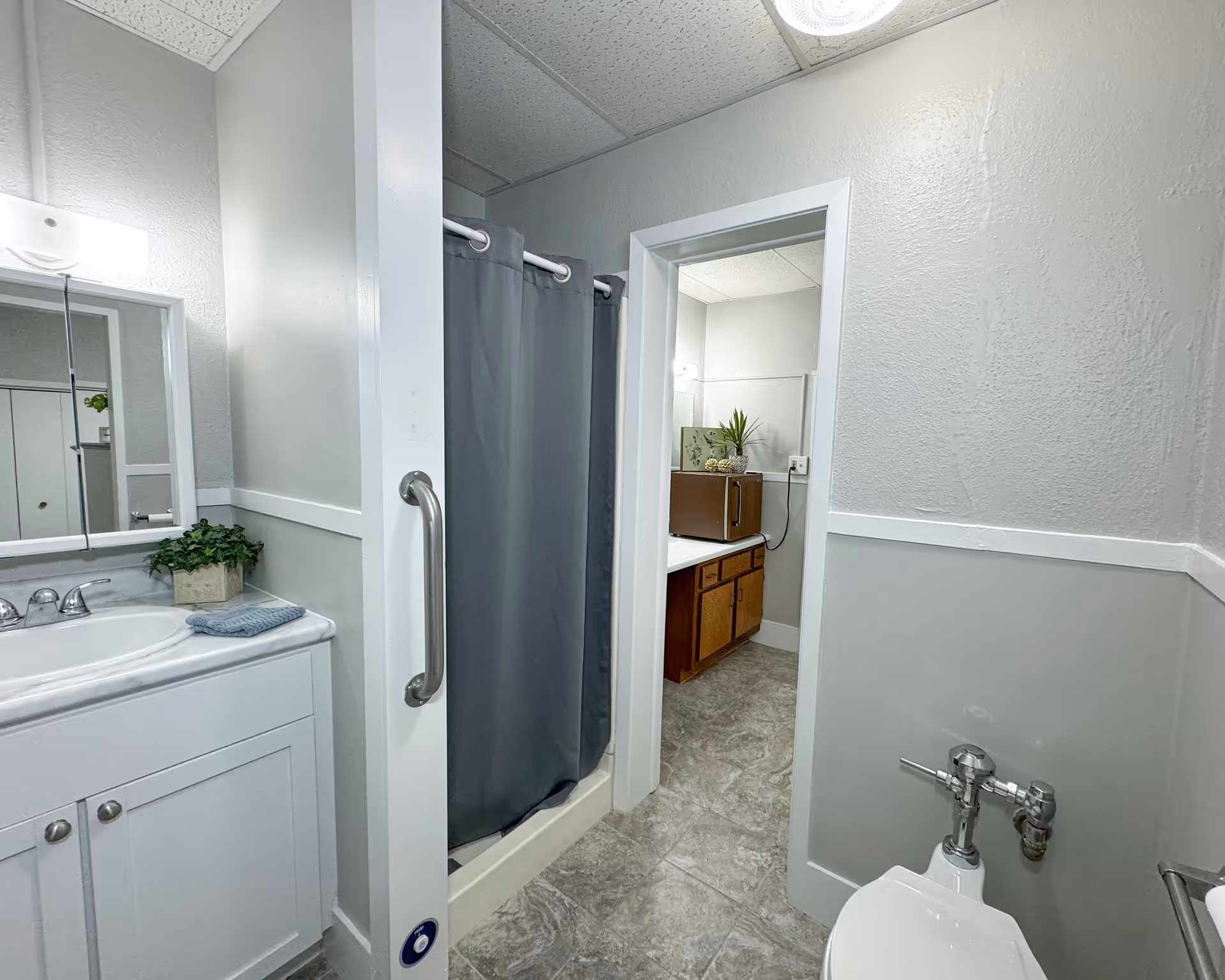 A bathroom with a white sink cabinet and mirror on the left, a shower with a gray curtain in the center, and a toilet on the right. Through an open doorway, a vanity with a wooden cabinet and a microwave on top is visible in an adjacent room. The walls are painted light gray with white trim, and the floor is covered with gray patterned tiles.
