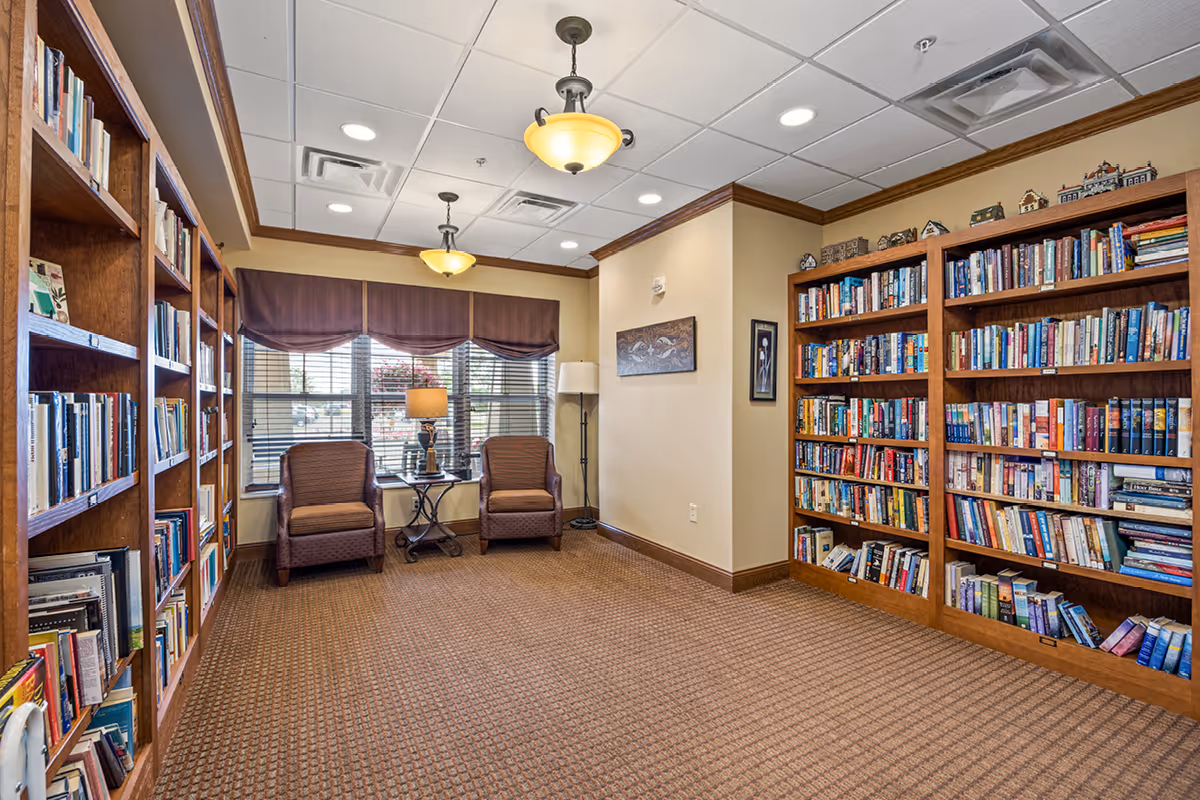 A cozy library/reading room with bookshelves lining the walls, two armchairs by a window, and warm ceiling lights.