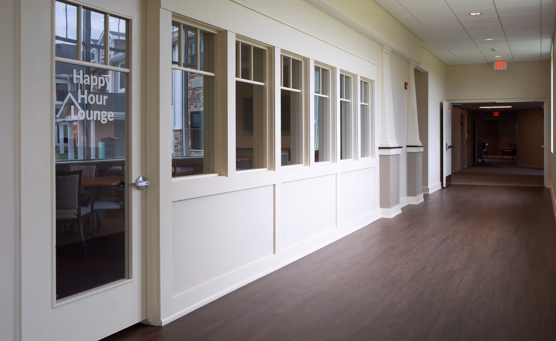 Empty interior hallway in a senior living facility with a door labeled "Happy Hour Lounge" and windows into the lounge.