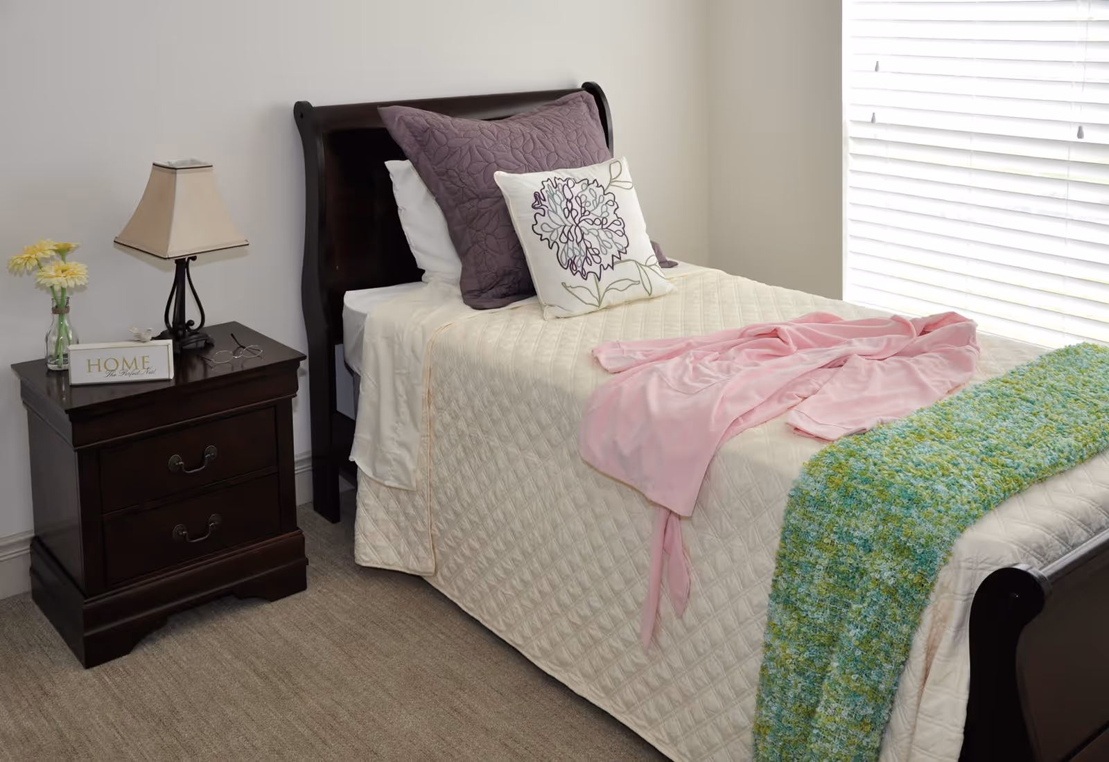 A neatly made single bed with a dark wooden frame, cream quilted bedspread, purple and floral pillows, a pink blanket, and a green knitted throw. Next to the bed is a dark wooden nightstand with a lamp, a small vase with yellow flowers, a pair of glasses, and a decorative sign that says 'HOME The Perfect Nest'. A window with white blinds is on the right side of the bed.