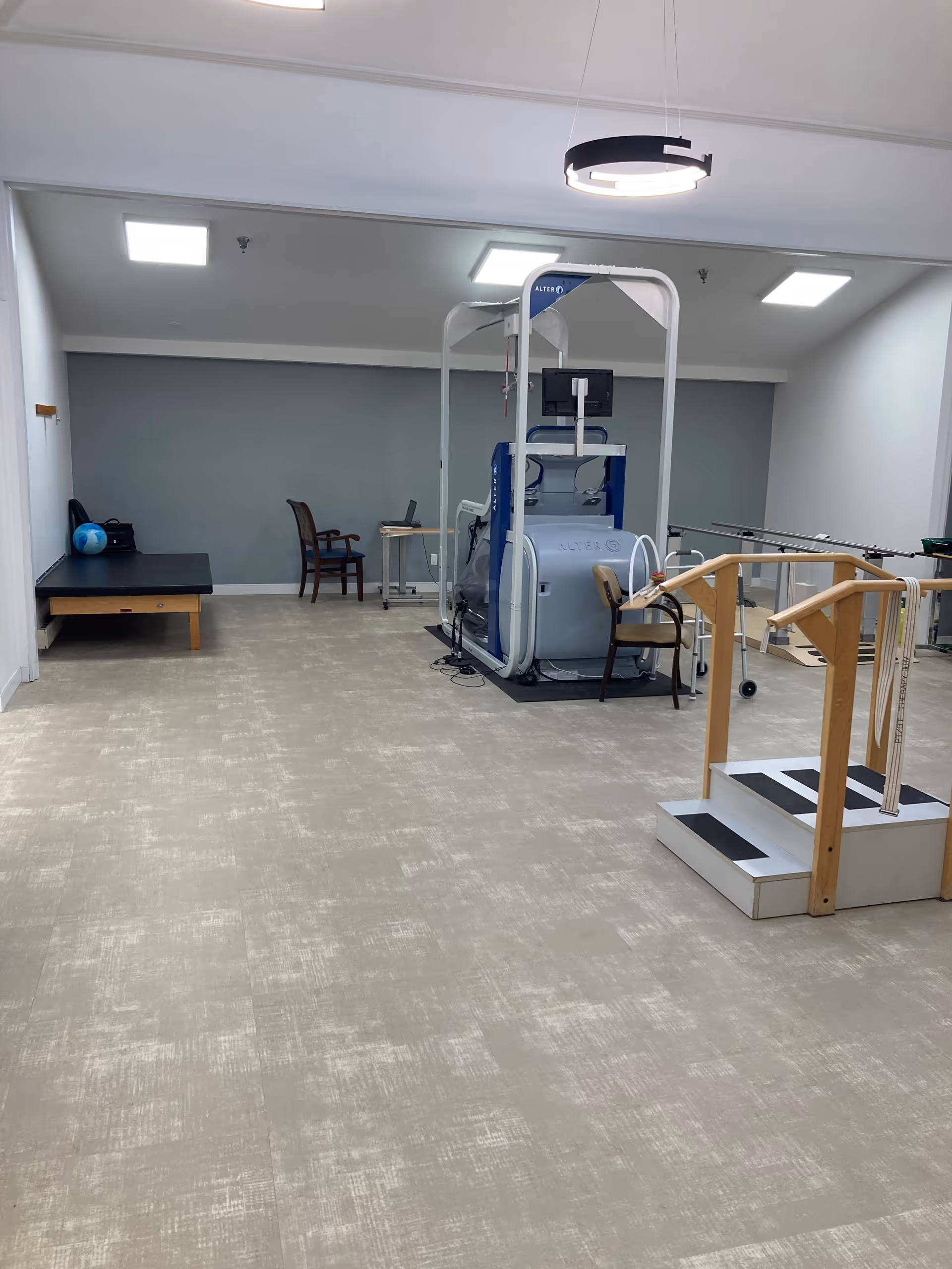 A rehabilitation or physical therapy room with exercise equipment including a treadmill-like machine, parallel bars for walking practice, a small set of stairs with handrails, a treatment table with a blue exercise ball on it, and a couple of chairs and a small table against the far wall. The room has a light-colored floor, white walls, and ceiling lights.