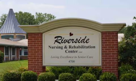 Brick sign reads Riverside Nursing and Rehabilitation Center with shrubs and lawn.