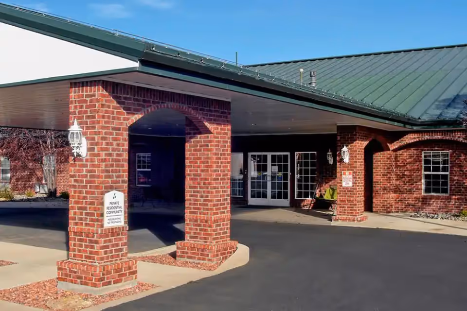 Entrance of a senior living facility with brick pillars supporting a green metal roof overhang, double glass doors, and a paved driveway. There is a sign on one of the pillars indicating it is a private residential community with no soliciting or trespassing.