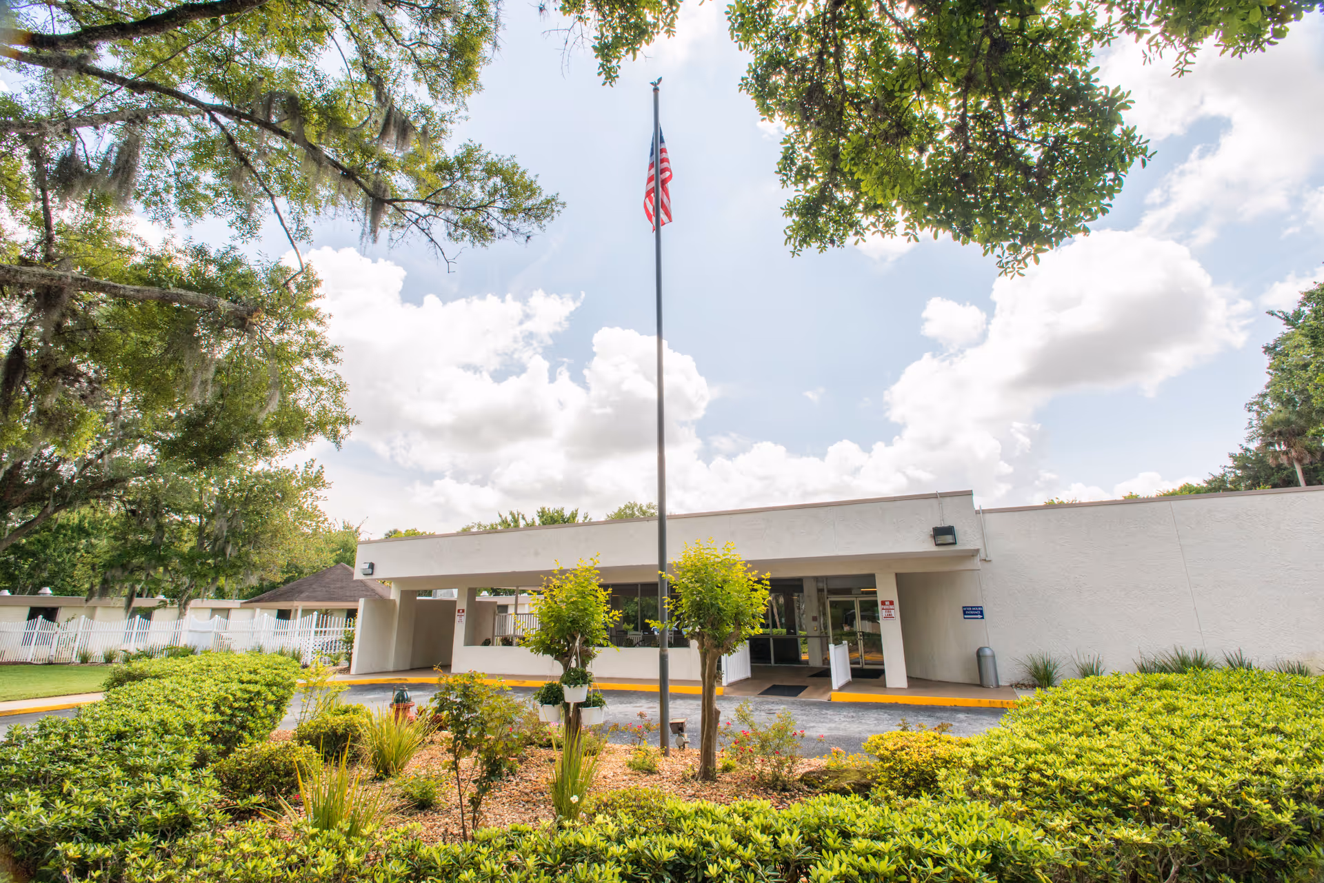 Exterior view of Daytona Beach Health and Rehabilitation Center showing a single-story building with a covered entrance. In front of the building is a flagpole with an American flag, surrounded by landscaped bushes and small trees. The sky is partly cloudy with sunlight filtering through the trees.