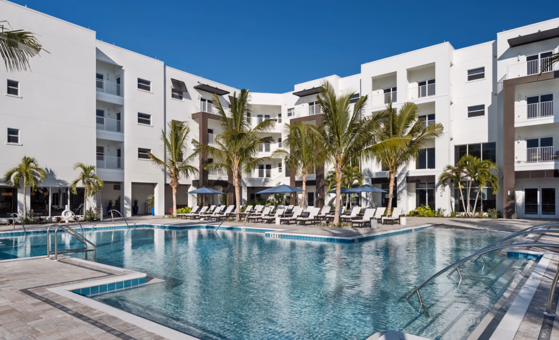 Outdoor swimming pool area at Amavida Senior Living - The Vistas Assisted Living with lounge chairs, palm trees, and a modern white multi-story building in the background under a clear blue sky.