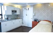 A room with a single bed covered with a beige quilt, a red bedside table with a telephone and other small items, white cabinets and a microwave mounted above a countertop with a mosaic tile backsplash, and a closed white door. The walls have a patterned wallpaper and the floor is dark wood.