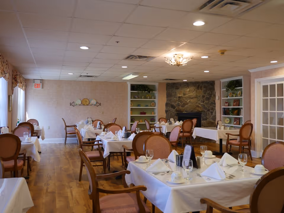 A dining room with multiple tables set with white tablecloths, chairs, place settings, and a stone fireplace in the background.