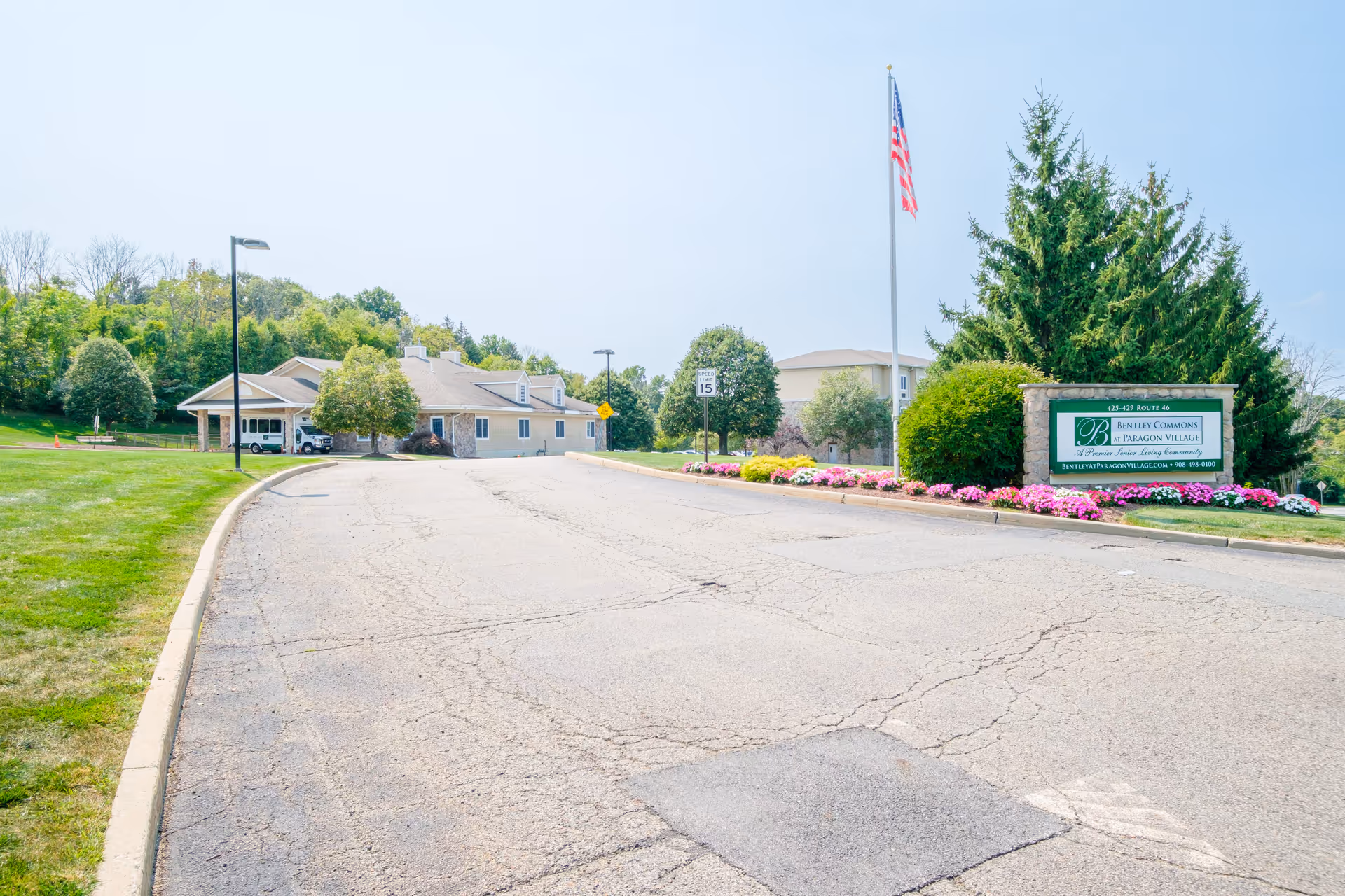 Driveway entrance to Bentley Commons at Paragon Village with the facility building, flagpole, landscaping and a stone sign by flowerbeds.