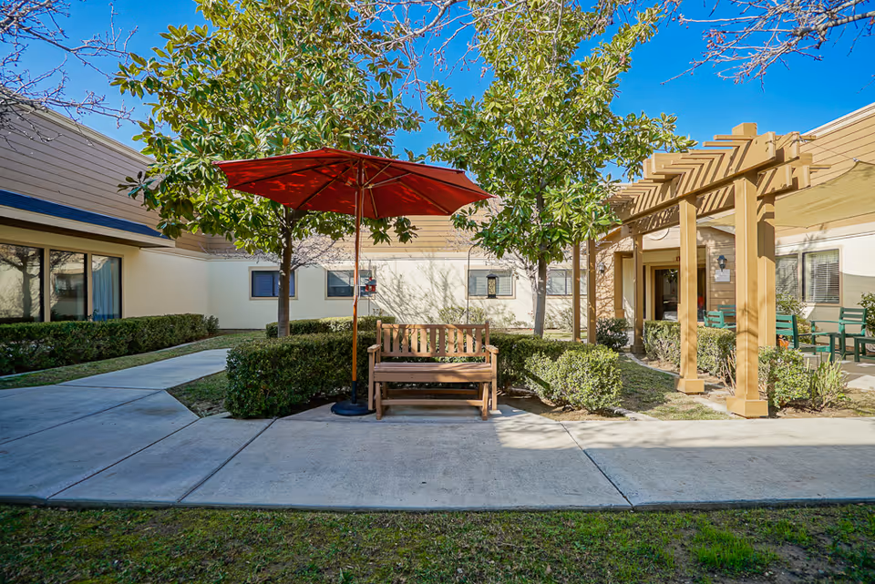 Outdoor courtyard area at Summerfield of Redlands featuring a wooden bench under a red umbrella, surrounded by green bushes and trees. There is a pergola structure to the right with additional seating, and the building walls with windows are visible in the background under a clear blue sky.