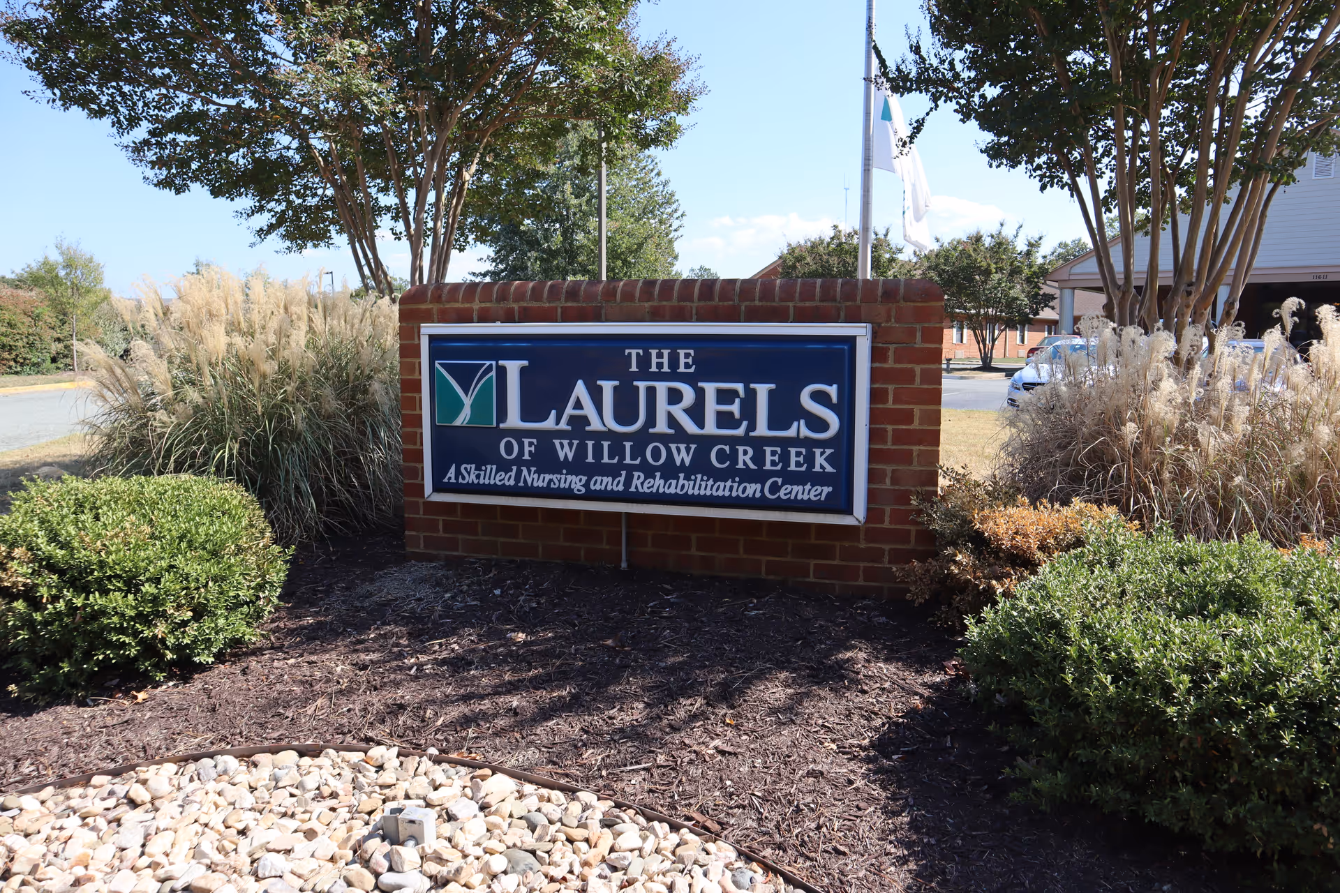 Outdoor view of a brick sign for The Laurels of Willow Creek, a skilled nursing and rehabilitation center, surrounded by bushes, ornamental grasses, and trees under a clear sky.