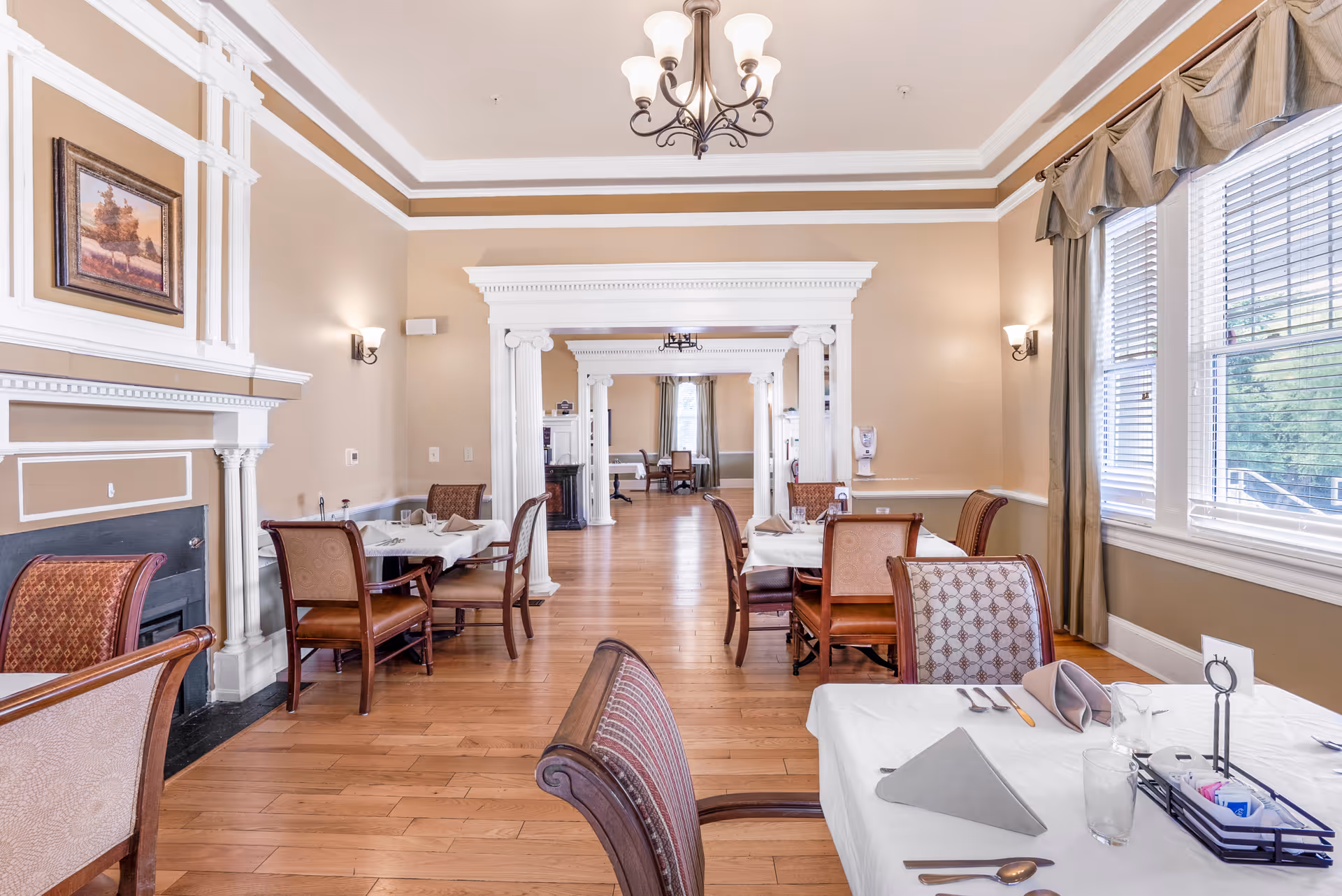 A bright and elegant dining room with wooden floors, beige walls, and white trim. Several tables are set with white tablecloths, napkins, glasses, and silverware. The room features a decorative fireplace with a framed painting above it, large windows with curtains allowing natural light, and a chandelier hanging from the ceiling. The room extends into another similarly styled room through a wide doorway with white columns.
