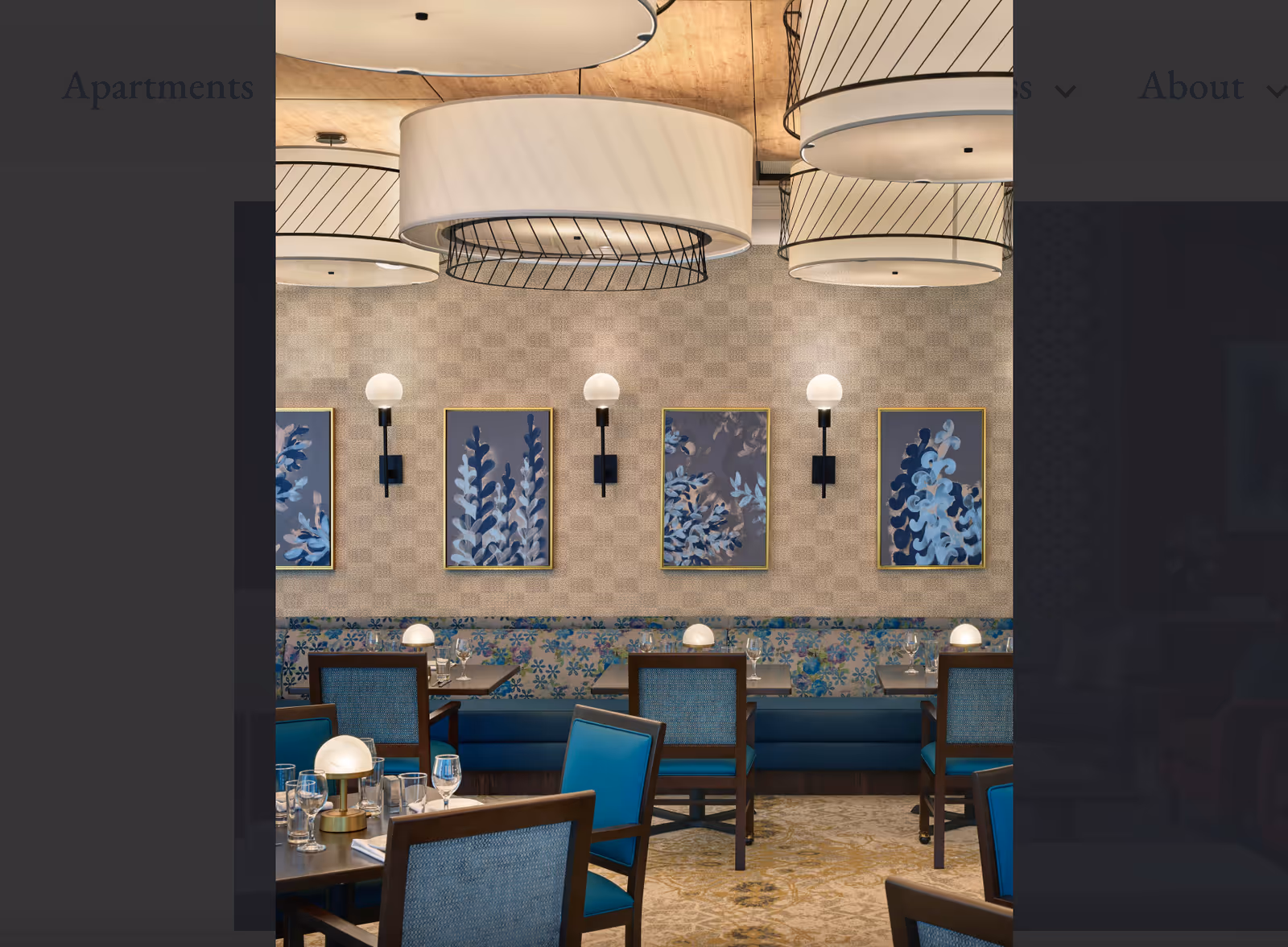 Dining room with blue upholstered chairs and banquette seating, round hanging light fixtures, framed botanical artwork, and set tables.