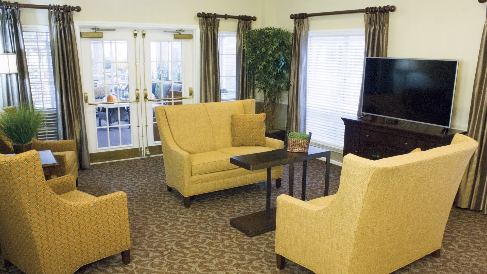 A cozy living room with yellow upholstered chairs and a loveseat arranged around a small dark wooden table. The room features large windows with brown curtains, a flat-screen TV on a dark wooden stand, and a glass door leading to an outdoor patio area. There are also decorative plants in the corners.