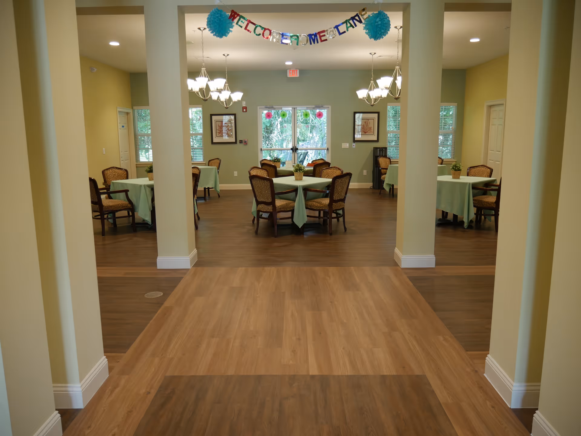 Interior view of a dining room in a senior living facility with several round tables covered with light green tablecloths and surrounded by wooden chairs with cushioned seats. The room has light yellow walls, wooden flooring, two chandeliers, framed artwork on the walls, and large windows letting in natural light. A colorful banner reading 'WELCOME FROME CAN' hangs across the entrance.