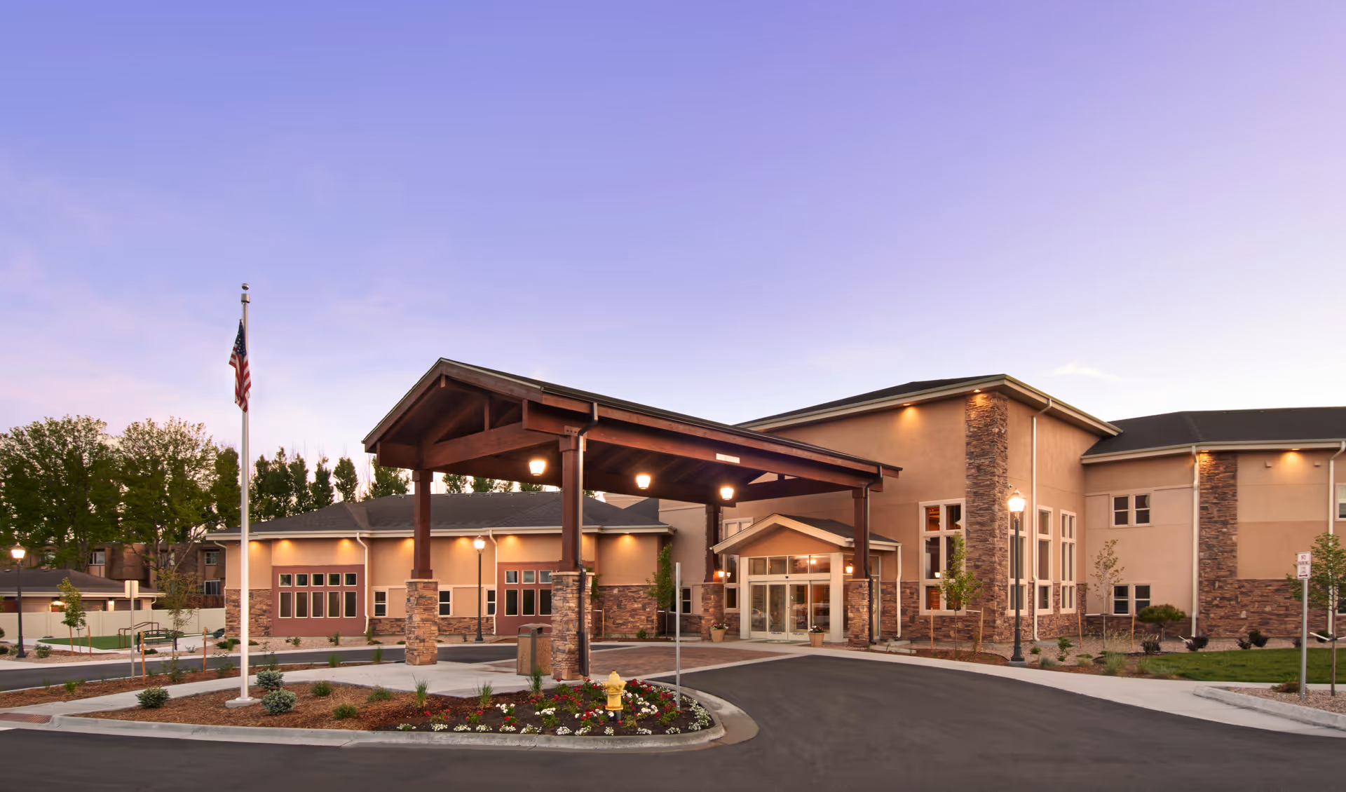 Exterior view of the Gardens on Quail senior living facility at dusk, featuring a covered entrance with stone pillars, landscaped flower beds, an American flag on a flagpole, and warm exterior lighting.