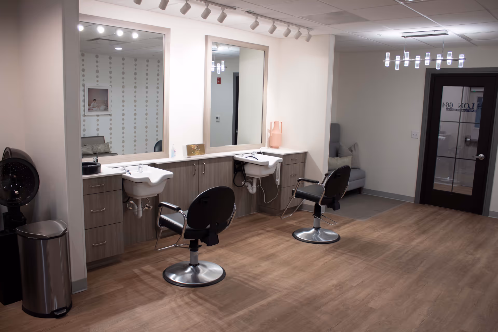 Interior view of a hair salon area in an assisted living facility featuring two salon chairs in front of two sinks with large mirrors above them. The room has wood flooring, light-colored walls, a modern chandelier, a gray armchair with pillows in the corner, and a door with glass panels.