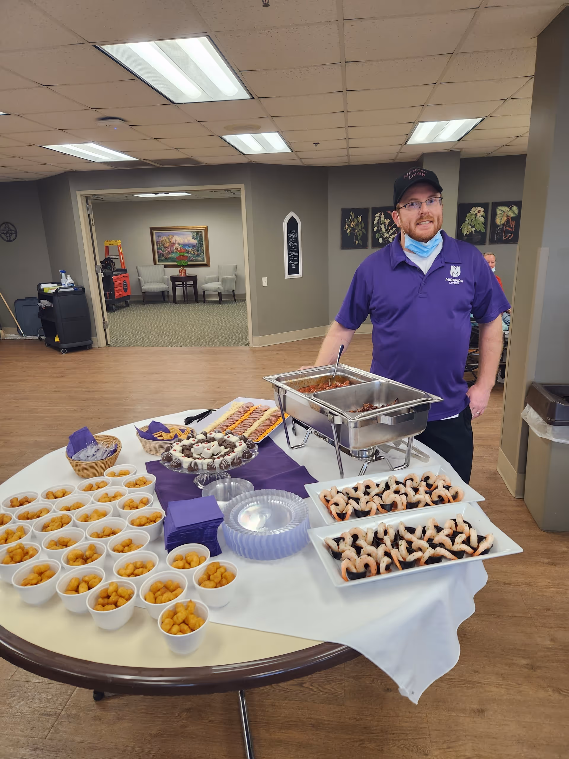 A staff member stands beside a buffet table laid out with shrimp, snack cups, and desserts in a senior living community common room.