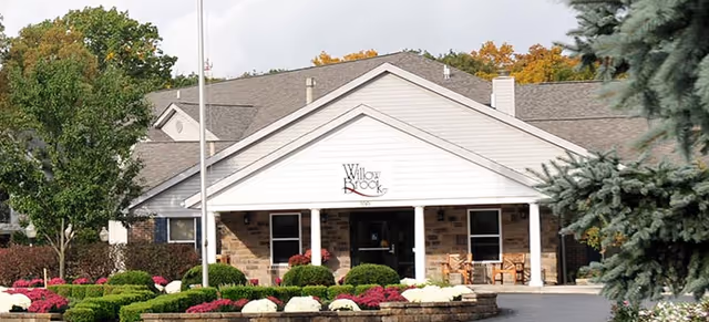 Front exterior view of Willow Brook Christian Village building with a peaked roof, white siding, brick lower walls, a flagpole, and landscaped bushes and flowers in front.