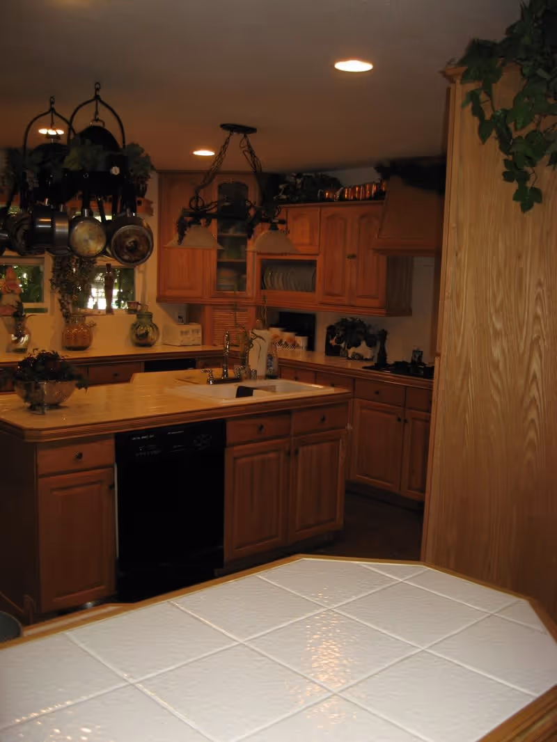 Kitchen with wooden cabinetry, a central island with sink and hanging pots, and a tiled countertop in the foreground.