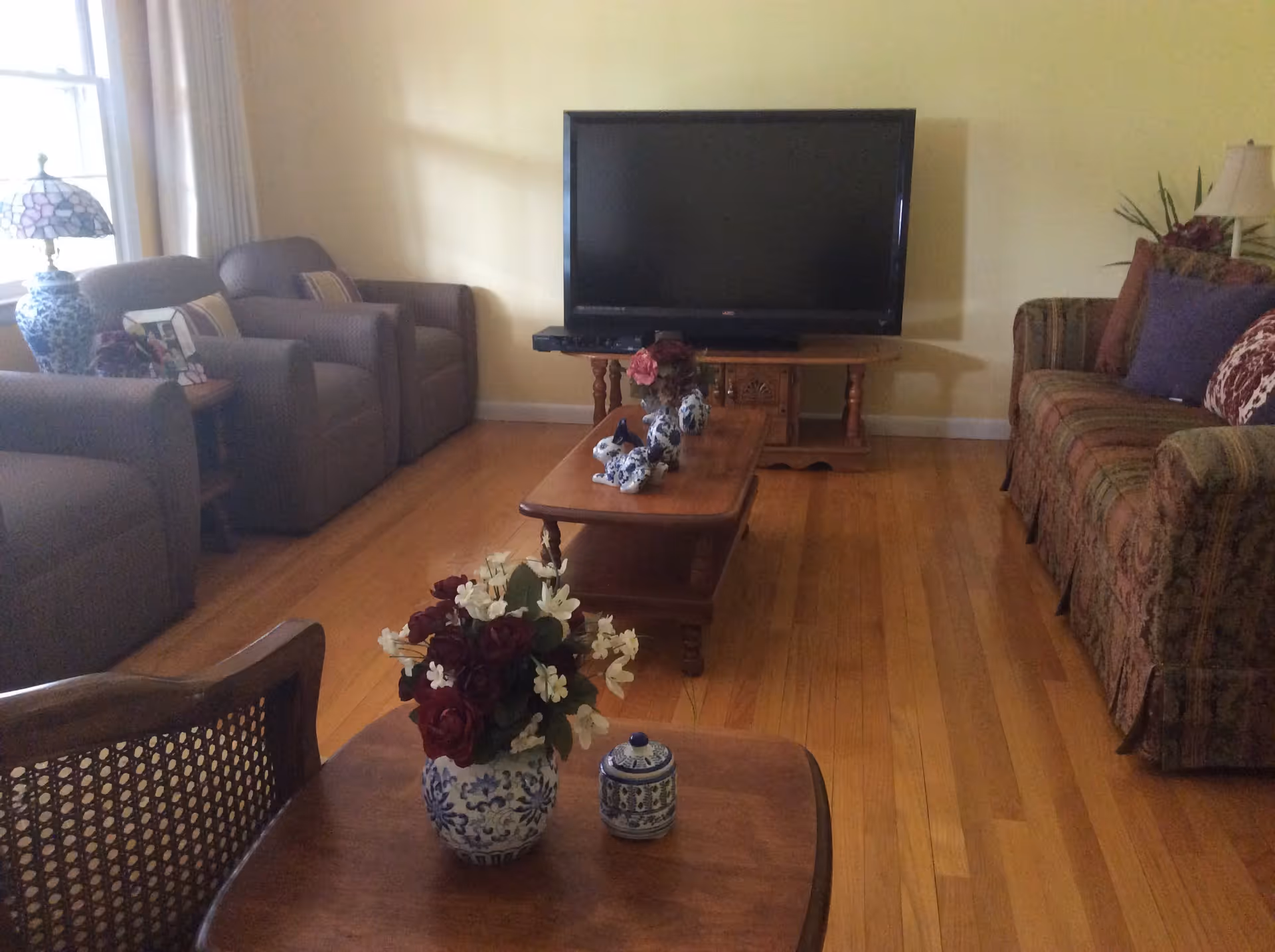A cozy living room with wooden flooring, featuring a large flat-screen TV on a wooden stand against a yellow wall. The room has a patterned sofa with multiple cushions on the right, two armchairs with pillows on the left near a window with white curtains, and a wooden coffee table in the center decorated with blue and white ceramic figurines and a flower arrangement. A side table with a blue and white lamp and another flower arrangement is also visible.