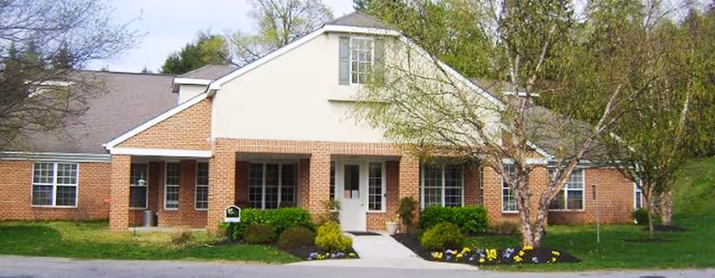 Front exterior view of a single-story brick building with a white entrance door, multiple windows, and a small garden with shrubs and flowers in front. Trees are visible on both sides of the building.